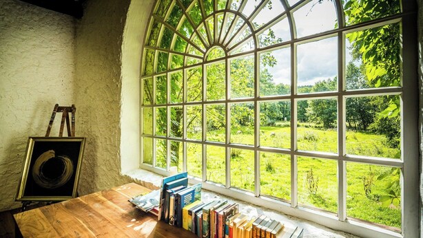 Ein Zimmer mit einem großen Bogenfenster, einem Holztisch mit Büchern und einem gerahmten Bild an der Wand.