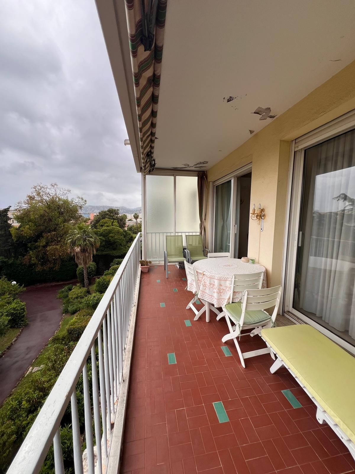 Ein Balkon mit Tisch und Stühlen, eine Schiebeglastür und Blick auf Grünflächen und eine Straße.