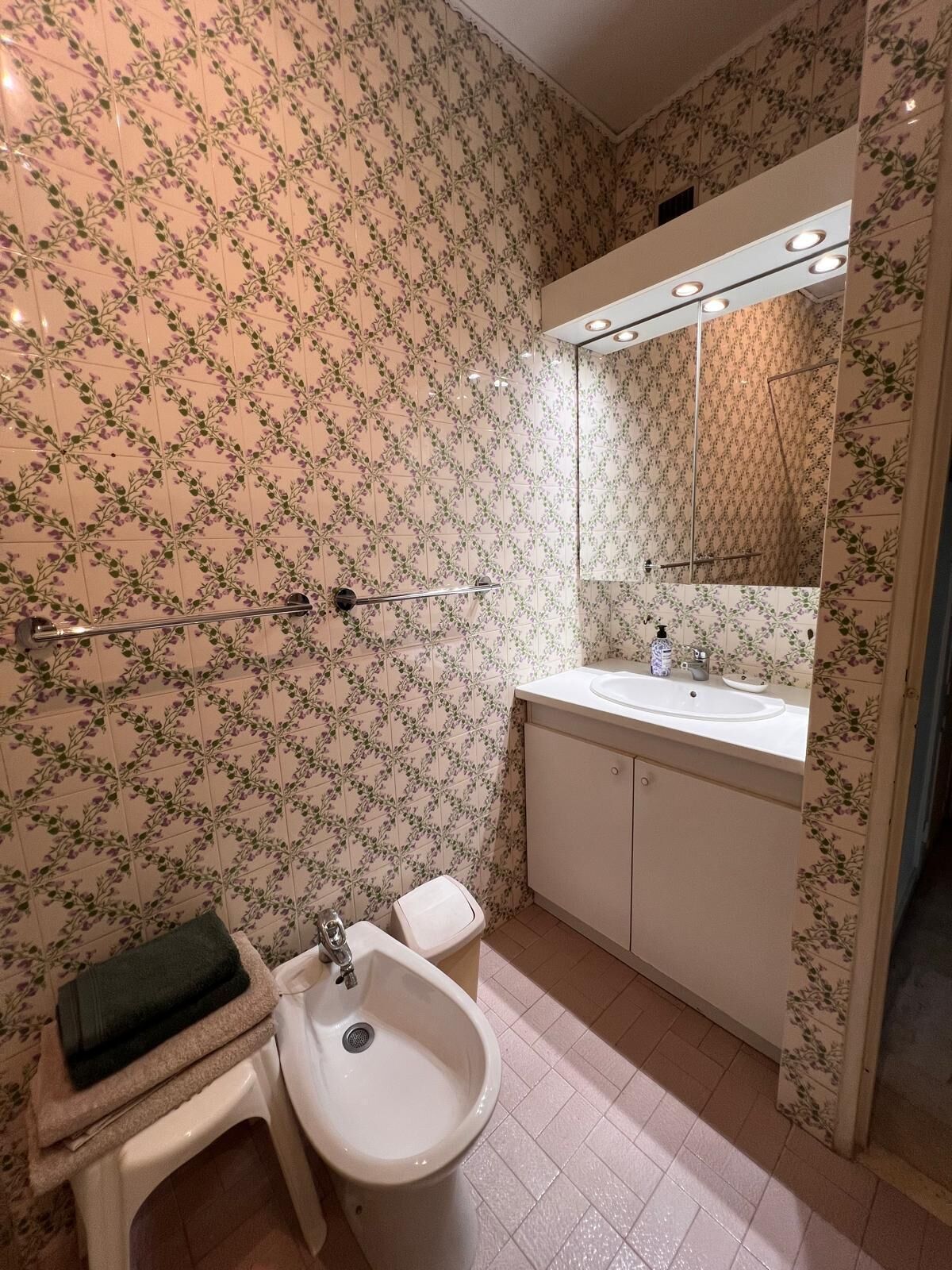 Un baño con papel tapiz floral, un lavabo blanco, un bidé y un espejo.