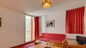 A cozy room with a red sofa, a wooden table, a TV, and a window with red curtains. - Central Slope Stay for Four (Les Deux Alpes)