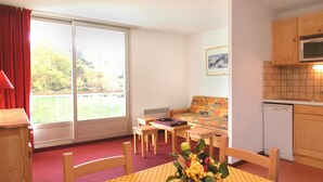 A room with a dining table, chairs, a sofa, a window with curtains, a kitchen area, and a small plant on the table. - Central Slope Stay for Four (Les Deux Alpes)