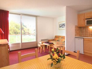 Ein Zimmer mit Esstisch, Stühlen, einer Couch, einem Fenster mit Vorhängen, einer Küchenzeile und einer kleinen Pflanze auf dem Tisch.