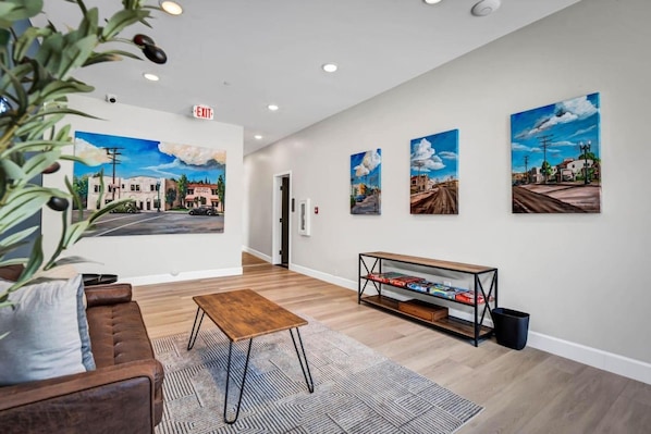 A hotel lobby with a brown leather sofa, a wooden coffee table, and a bookshelf. There are four paintings on the wall depicting urban scenes.