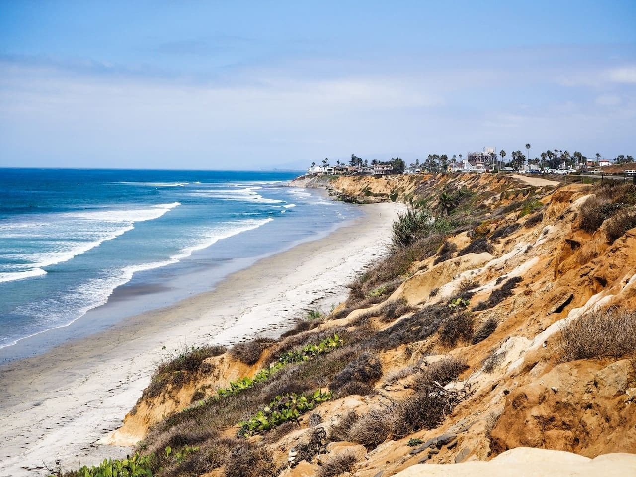A coastal landscape with a sandy beach, rocky cliffs, and a distant shoreline with buildings.