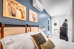 Habitación de hotel con una cama, dos cuadros en la pared, una lámpara de noche y una puerta que da a un pasillo.