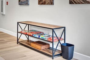 A wooden console table with metal frame shelves, holding board games and a storage box, against a plain wall with a white baseboard.