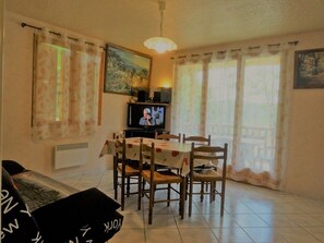 A living room with a dining table, chairs, a sofa, a television, and curtains. - Gite les Acacias lesgitesdeshautsdevialot.fr (Auriac-du-Périgord)