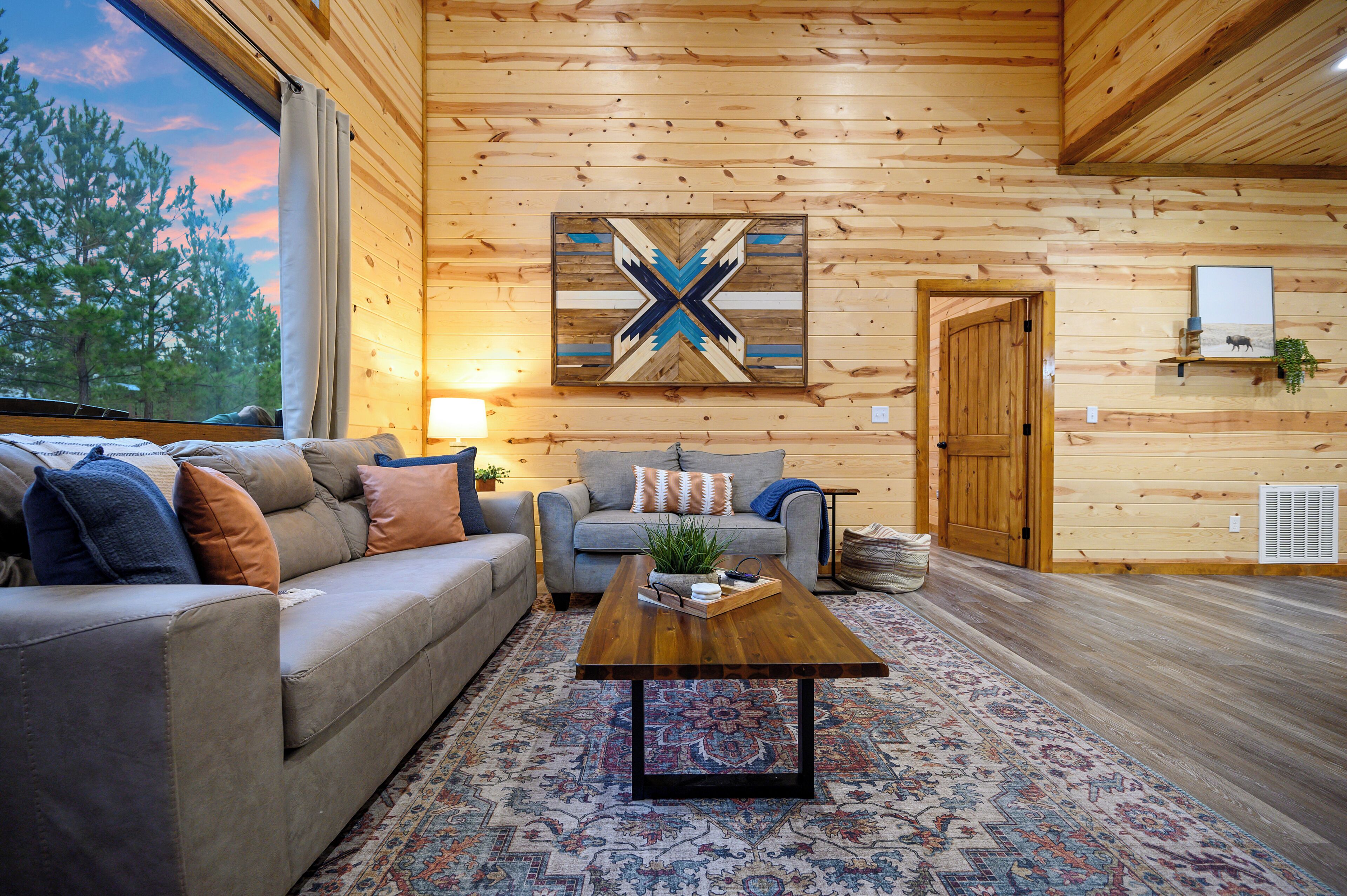 Ein gemütliches Blockhaus-Interieur mit Holzbalkendecke, einem großen Fenster, einer Couch, einem Couchtisch und einem gemusterten Teppich.