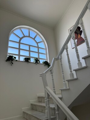 Un escalier avec des balustrades blanches, un palier en marbre et une grande fenêtre en arc surmontée d’un rebord où sont disposées des plantes.