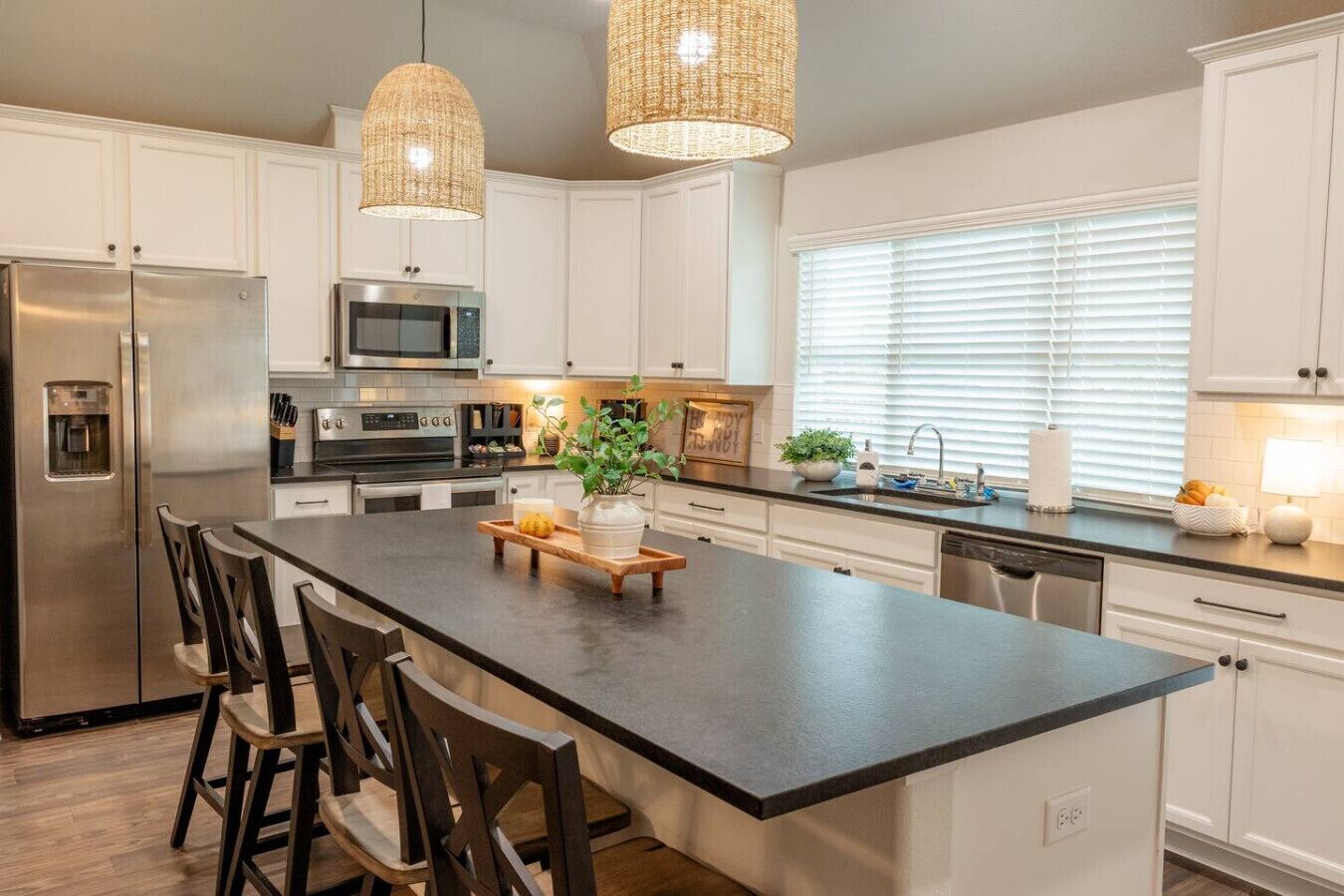A modern kitchen with white cabinets, stainless steel appliances, a large island with bar stools, and wicker pendant lights.