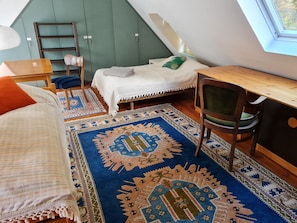 A loft bedroom with a sloped ceiling, a bed with a white bedspread, a desk with a chair, and a large window.