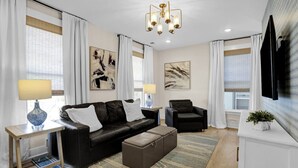 A modern living room with a black leather sofa, a matching ottoman, a side table with a lamp, a black armchair, and a television mounted on the wall. - Walk to Memphis Nightlife | 100 Yr Old Cozy Apt (Memphis)