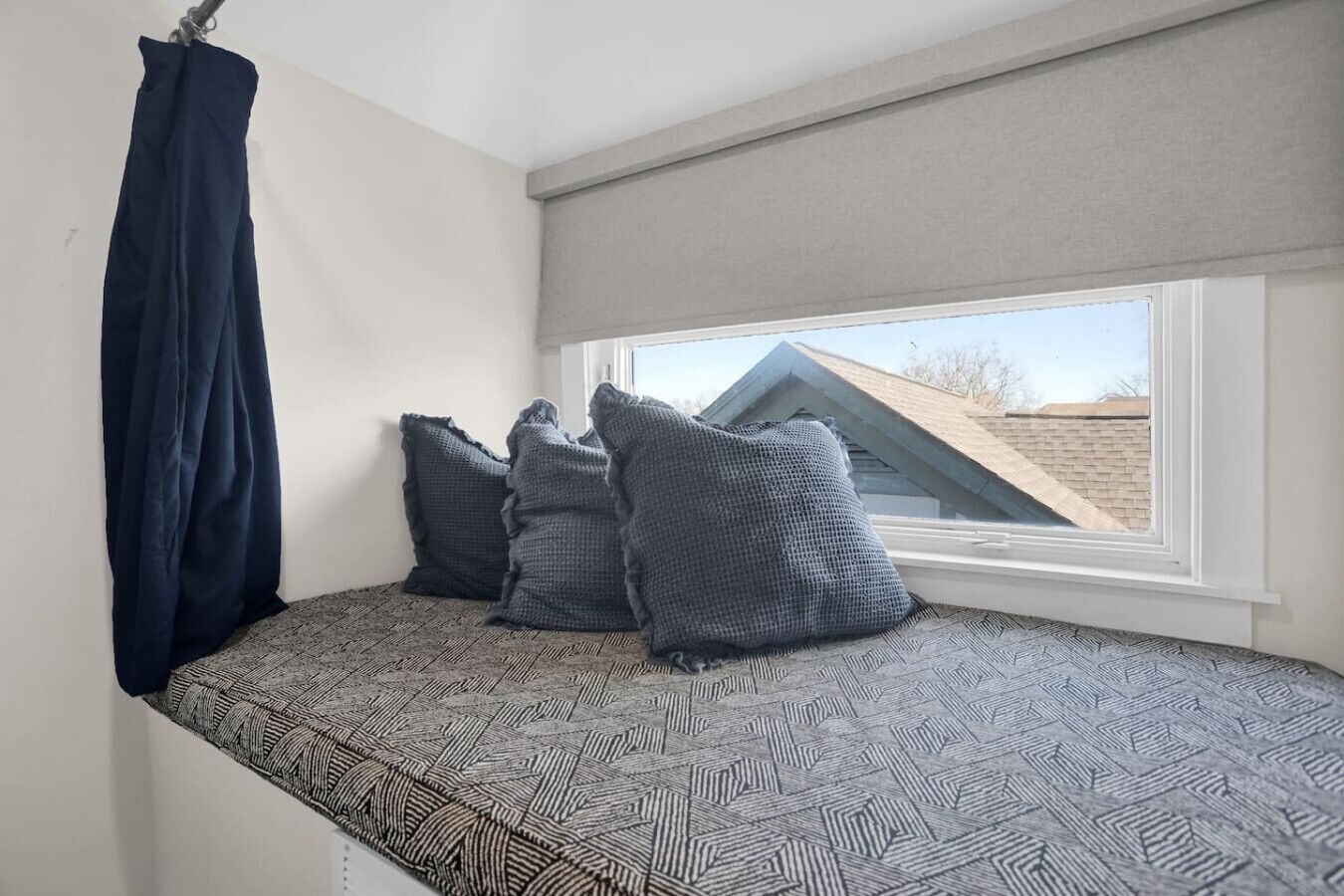 Een klein slaapverblijf met een raam, een gestreept matras en twee kussens, een donker gordijn, en uitzicht op een gebouw.