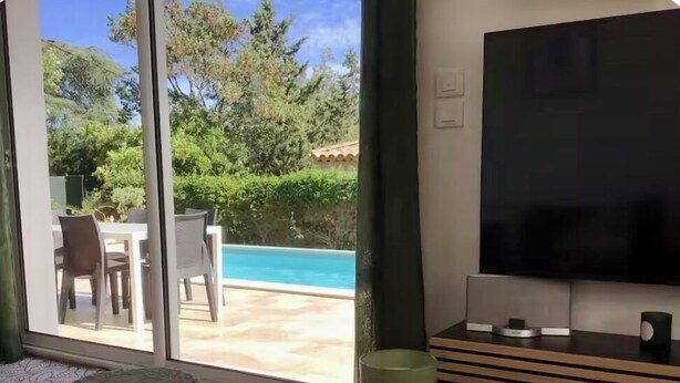 Una sala de estar moderna con televisor, una mesita redonda y una zona de estar con vista a una piscina y vegetación.