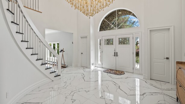 A grand foyer with a marble floor, a staircase, a chandelier, and a large arched window.