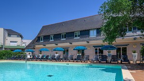A swimming pool with lounge chairs and umbrellas, a building with a sloped roof, and a clear blue sky. - Studio With Balcony Near Vannes (Plescop)