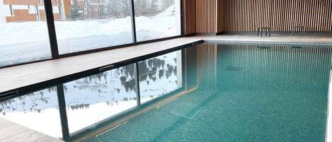 Piscina interior transparente con vista a un paisaje nevado a través de amplios ventanales.