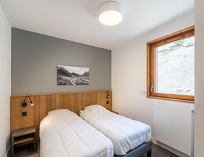 Ein Schlafzimmer mit zwei Betten, einem Holzkopfende, einem Nachttisch, einer Wandlampe, einem Fenster mit Blick auf eine Berglandschaft und einer Deckenleuchte.