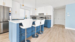 A modern kitchen with a stainless steel refrigerator, white cabinets, blue bar stools, and a tiled backsplash.