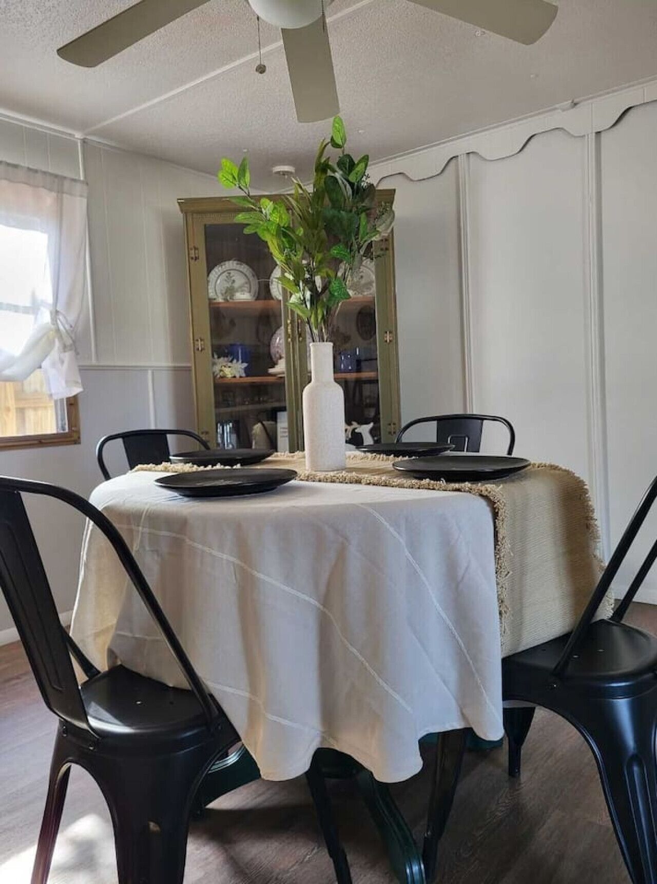 A dining area with a table set for four, surrounded by black chairs. A white tablecloth covers the table, and a white vase with green plants is placed in the center. Behind the table, there is a glass cabinet with decorative plates.