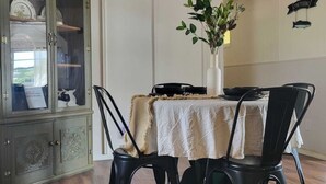 Een eetruimte met een tafel voor vier personen, omringd door zwarte stoelen, een witte tafelkleed en een vaas met groene planten. Links is een kast met glazen deuren zichtbaar.