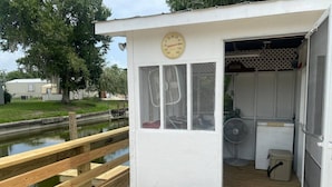 Een klein, wit tuinhuisje met een thermometer op de deur, een ventilator binnenin en een houten steiger die naar een vijver leidt.