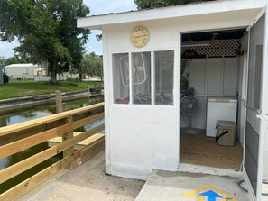 A small, white utility shed with a thermometer on the door, a fan inside, and a wooden dock leading to a pond. - Pet Friendly | Private Dock & Direct Canal Access (Okeechobee)