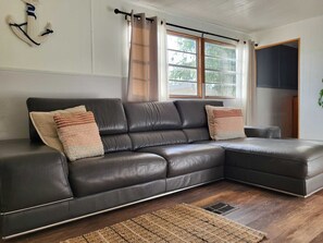 A dark leather sectional sofa with two cushions, a wooden floor, a window with curtains, and a plant on the right.