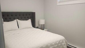 A bedroom with a bed, a nightstand, a lamp, and a window with blinds. - Go-HabitAt: Home away from Home (Saskatoon)