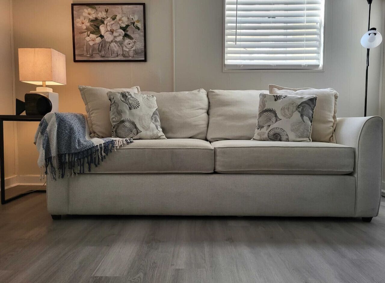 Un canapé beige agrémenté de coussins à motifs, un plaid bleu, une œuvre d’art florale encadrée et une lampe sur pied.