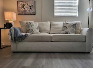 A beige sofa with patterned cushions, a blue throw blanket, a framed floral artwork, and a floor lamp.
