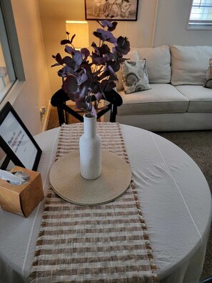 A round table with a woven placemat, a white vase with purple flowers, a framed picture, and a tissue box.