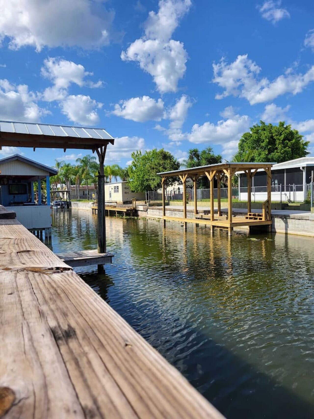 A wooden dock extending into calm water with a covered seating area and a building in the background.