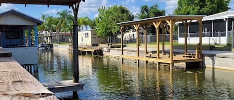 A wooden dock extending into calm water with a covered seating area and a building in the background.