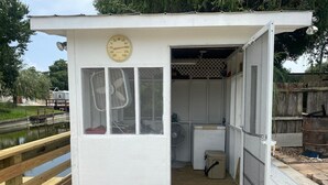 Ein kleines, weißes Nebengebäude mit einer Uhr an der Tür, einem Ventilator im Inneren und einem hölzernen Steg im Hintergrund.