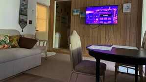 Ein Wohnzimmer mit einer Couch, einem Esstisch, einem an einer Holzwand montierten Fernseher und einer Tür zu einem anderen Raum.