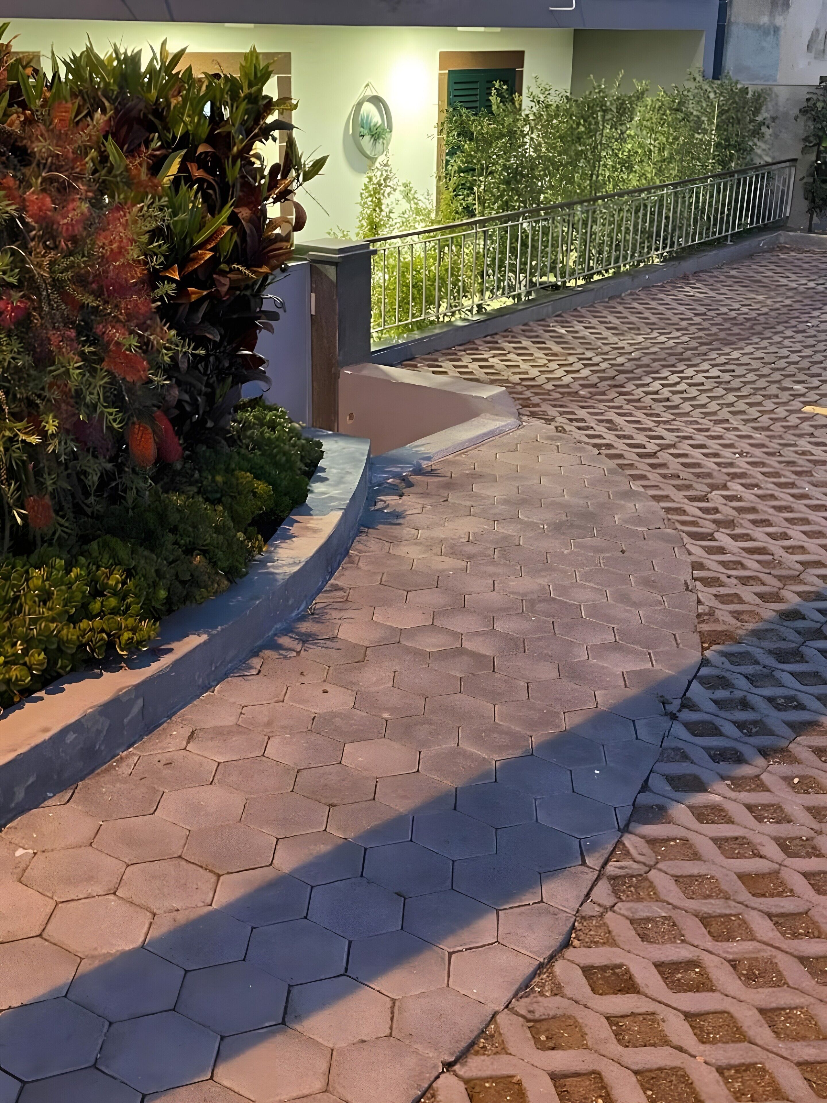 A paved pathway with hexagonal tiles, bordered by greenery and a metal railing.