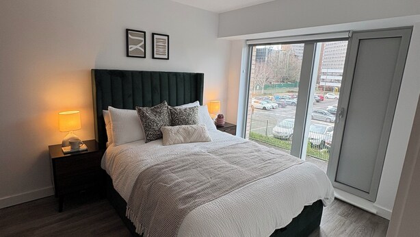 Een moderne slaapkamer met een groot bed, twee bedlampen, een donker hoofdbord en uitzicht op een parkeerplaats door een schuifdeur.