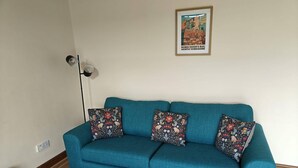 A teal sofa with floral-patterned cushions, a framed picture on the wall, and a floor lamp. - Lovely 3-bedroom house near Robin Hood's Bay perfect for relaxing peaceful break (Robin Hood's Bay)