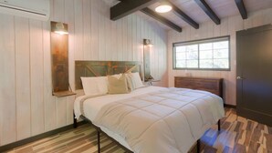 Ein Schlafzimmer mit hölzernem Kopfteil, einem Bett mit weißer Bettwäsche, einem Nachttisch aus Holz, einer Kommode und einem Fenster mit Blick.