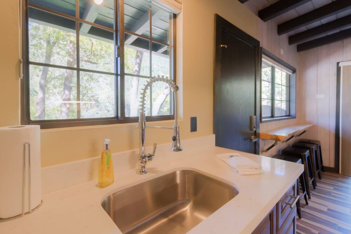 A modern kitchen with a stainless steel sink, a window with a view of trees, and a bar area with stools.