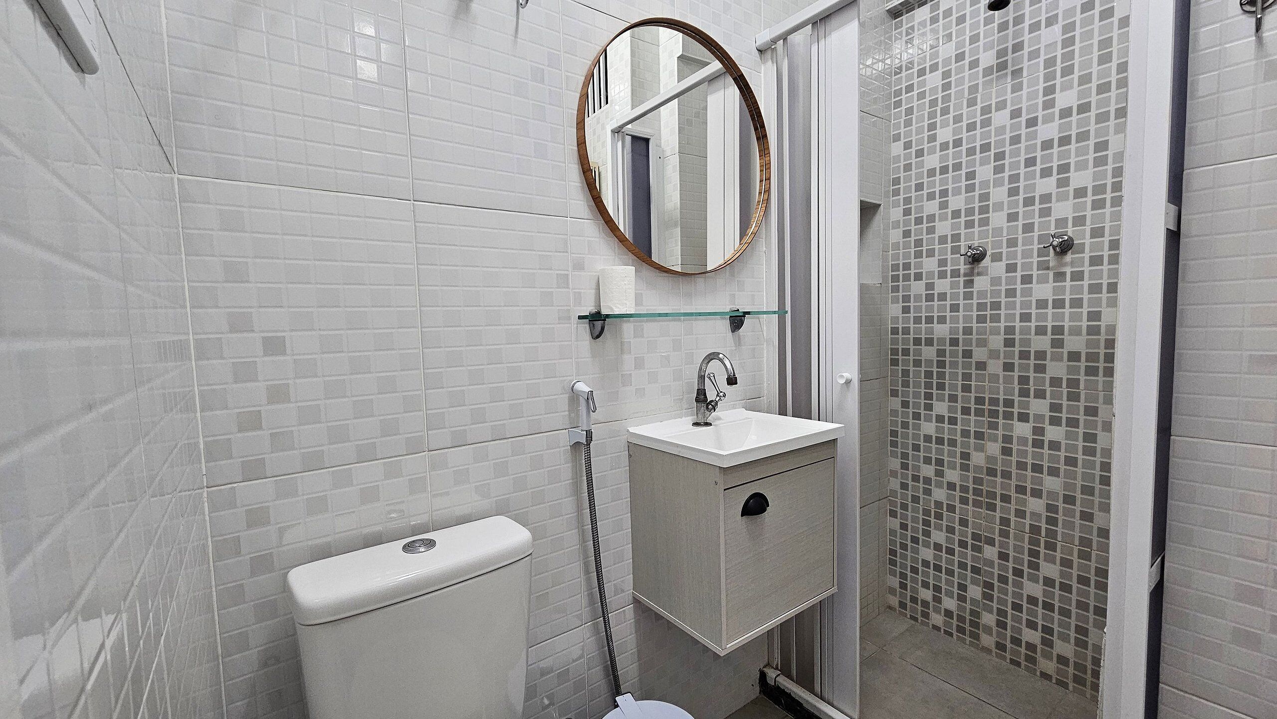 Een badkamer met witte betegelde muren, een glazen plank boven de wastafel, een toilet en een douche met mozaïektegels.