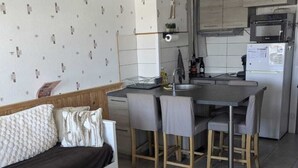 Ein kompakter Wohnbereich mit einer Couch, einem Esstisch mit Stühlen, einer Küchenzeile mit Mikrowelle und Kühlschrank sowie einer kleinen Holzbank.