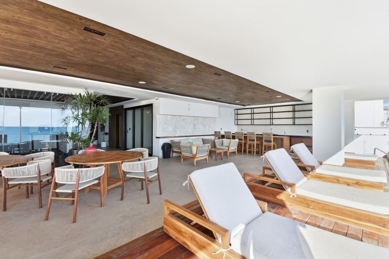 A spacious indoor area with a wooden ceiling, a large table with chairs, and several lounge chairs.