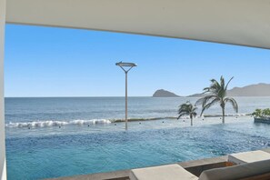 Blick auf ein Schwimmbecken mit freiem Blick auf das Meer, eine Palme und ein Berg in der Ferne.