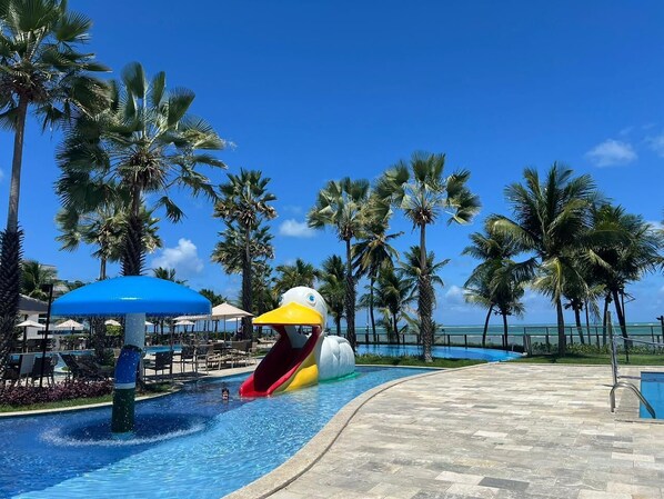 Une piscine avec un toboggan en forme de canard, des palmiers et un ciel bleu azur.