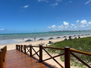 Ein hölzerner Steg führt zu einem Strand mit Sonnenschirmen und Liegestühlen.