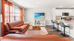 Una sala de estar moderna con un sofá, una mesa de centro y un televisor. Al lado, una cocina con gabinetes blancos, un microondas y una zona de bar con taburetes.