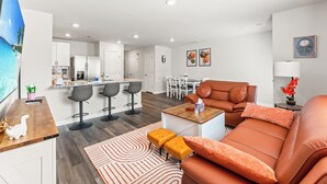 Una sala de estar moderna con sofá, reposapiés y mesa de centro, una cocina con armarios blancos y un comedor con mesa y sillas.
