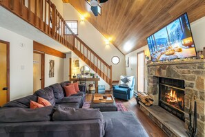 Ein Wohnzimmer mit Kamin aus Stein, einem großen Flachbildfernseher, einem dunkelgrauen Sofa und einer Holztreppe.